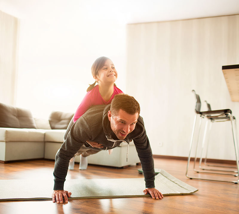 a dad does push-ups in the family room while his daughter sits on his back and laughs