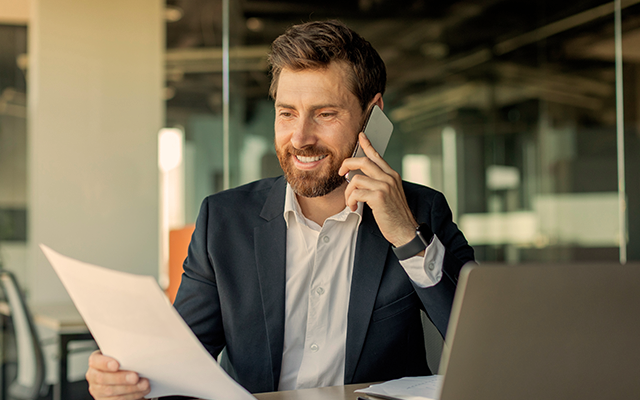 Man smiling holding papers while on the phone in an office.