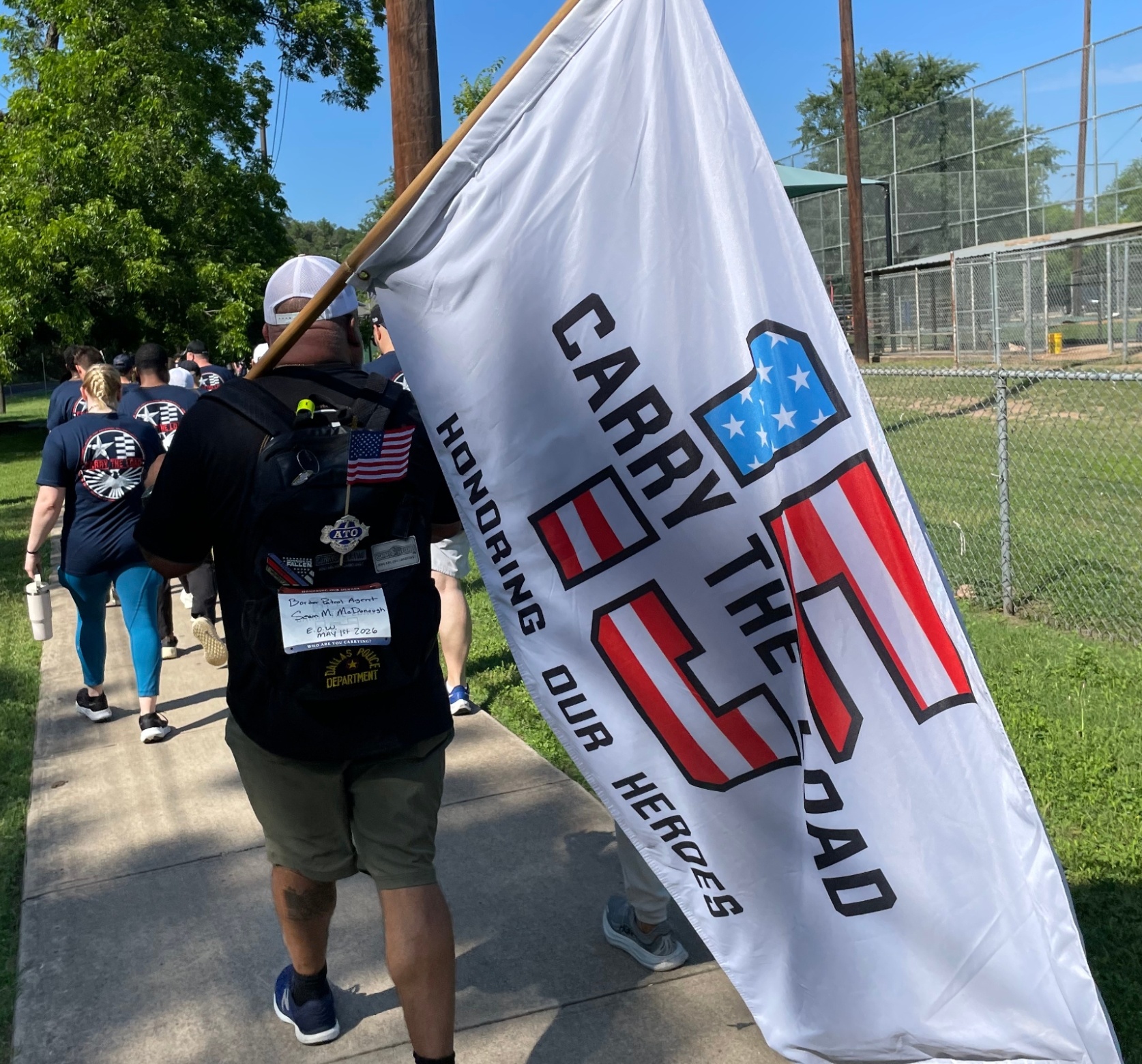 A G.E.H.A employee walks through a park carrying a white flag that reads "Carry the load, honoring our heroes" with a graphic of the number 15 in US flag print. 
